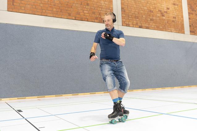 Mann mit Kopfh&ouml;rern skatet auf Rollschuhen in einer Sporthalle mit grauen W&auml;nden und Holzverkleidung.