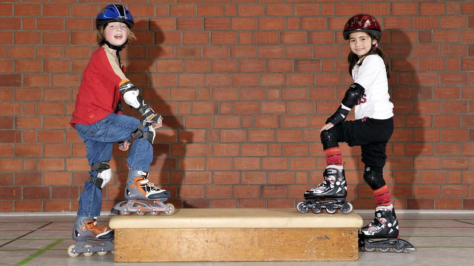 Zwei Kinder auf Rollskates stehen auf einer Holzplattform in einer Halle, beide tragen Helme und Schutzausrüstung.