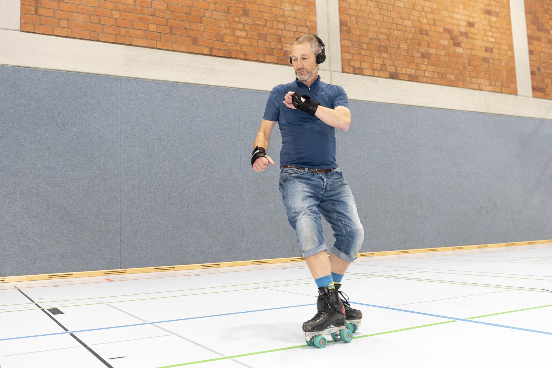Mann mit Kopfh&ouml;rern skatet auf Rollschuhen in einer Sporthalle mit grauen W&auml;nden und Holzverkleidung.