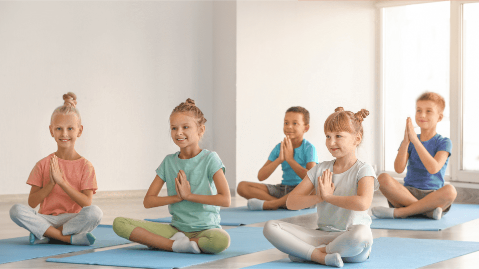 Fünf Kinder sitzen im Lotussitz auf blauen Yogamatten und halten die Hände in Anjali-Mudra vor der Brust.
