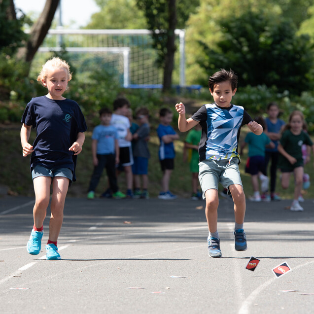 Zwei Kinder rennen auf einem Sportplatz, w&auml;hrend andere im Hintergrund zuschauen. Sonniger Tag, gr&uuml;ne B&auml;ume.