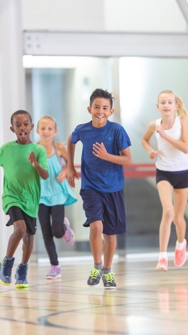Sechsäugige Kinder rennen freudig auf einem sportlichen Hallenboden in bunter Sportkleidung.