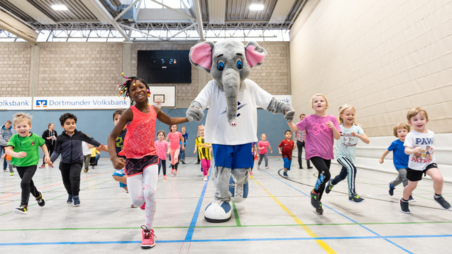 Kinder und ein Elefanten-Maskottchen laufen gemeinsam in einer Sporthalle w&auml;hrend einer Aktivit&auml;tsveranstaltung.