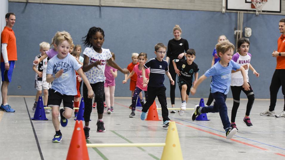 Kinder laufen in einer Sporthalle durch einen Slalomparcours aus Kegeln und springen über Hindernisse.