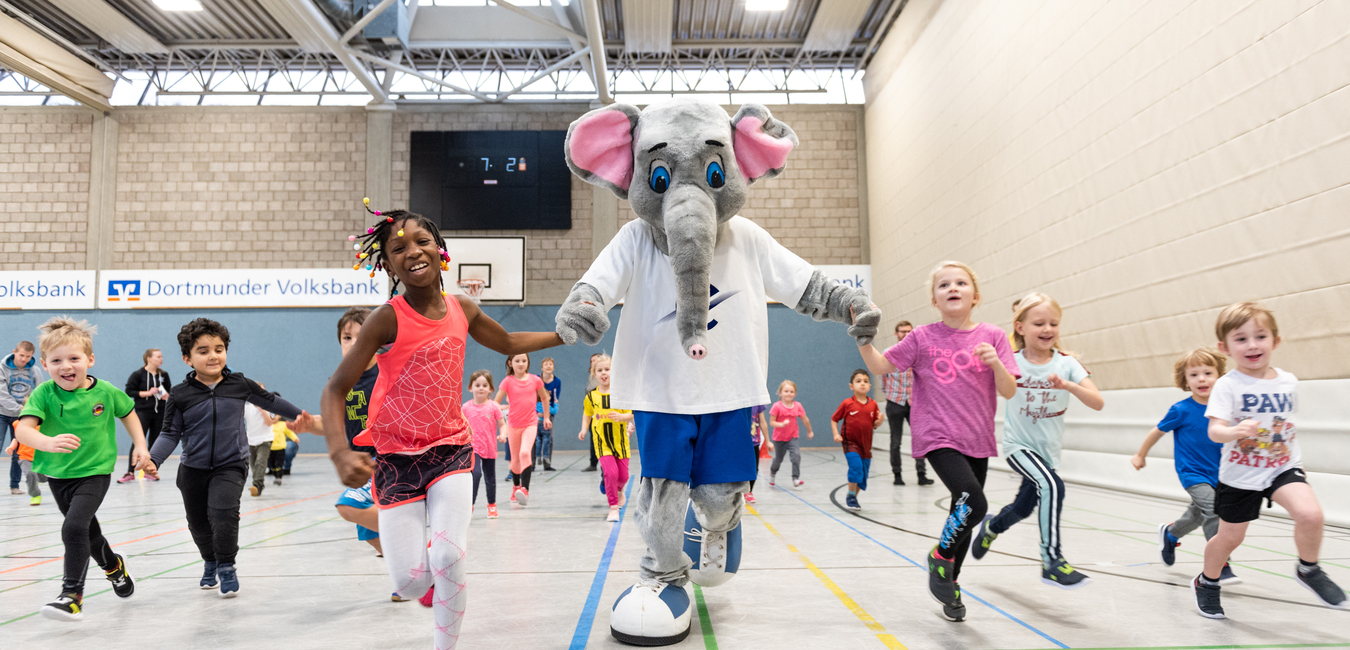 Kinder und ein Elefanten-Maskottchen laufen gemeinsam in einer Sporthalle w&auml;hrend einer Aktivit&auml;tsveranstaltung.