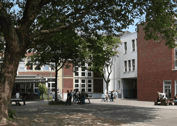Schülergruppen stehen im Schulhof, umgeben von Gebäuden und Bäumen, an einem sonnigen Tag.