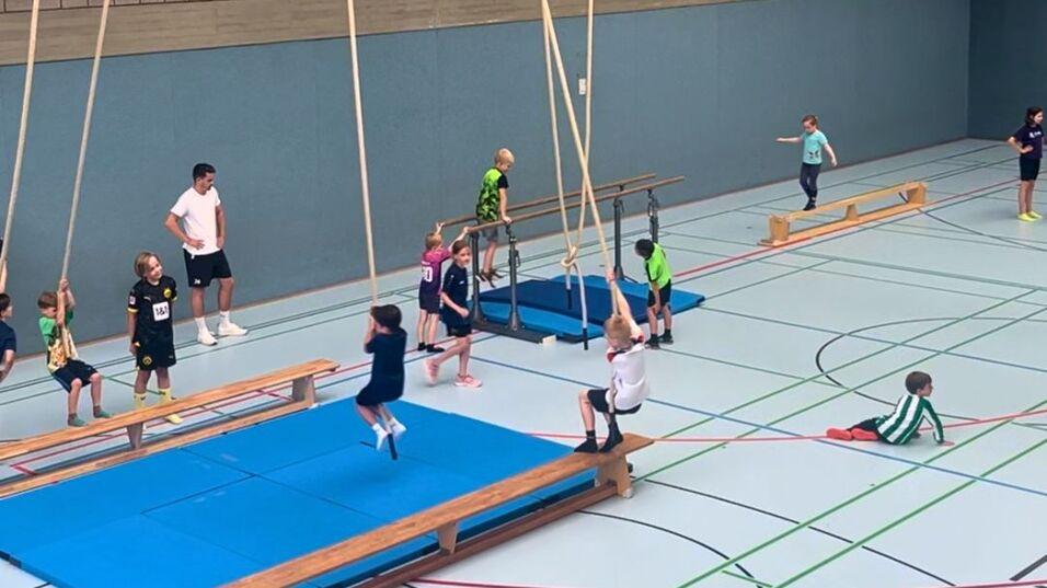Kinder trainieren in einer Sporthalle an verschiedenen Geräten, darunter Schwebebalken und Seile.