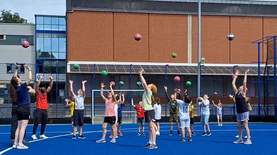 Gruppentraining auf einem blauen Sportplatz, Kinder werfen Bälle in die Luft, umringt von Erwachsenen.