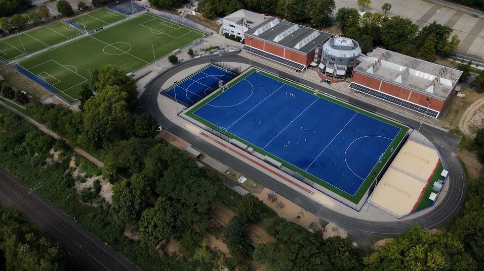 Luftaufnahme eines Sportkomplexes mit einem blauen Hockeyfeld und mehreren Fußballplätzen im Hintergrund.