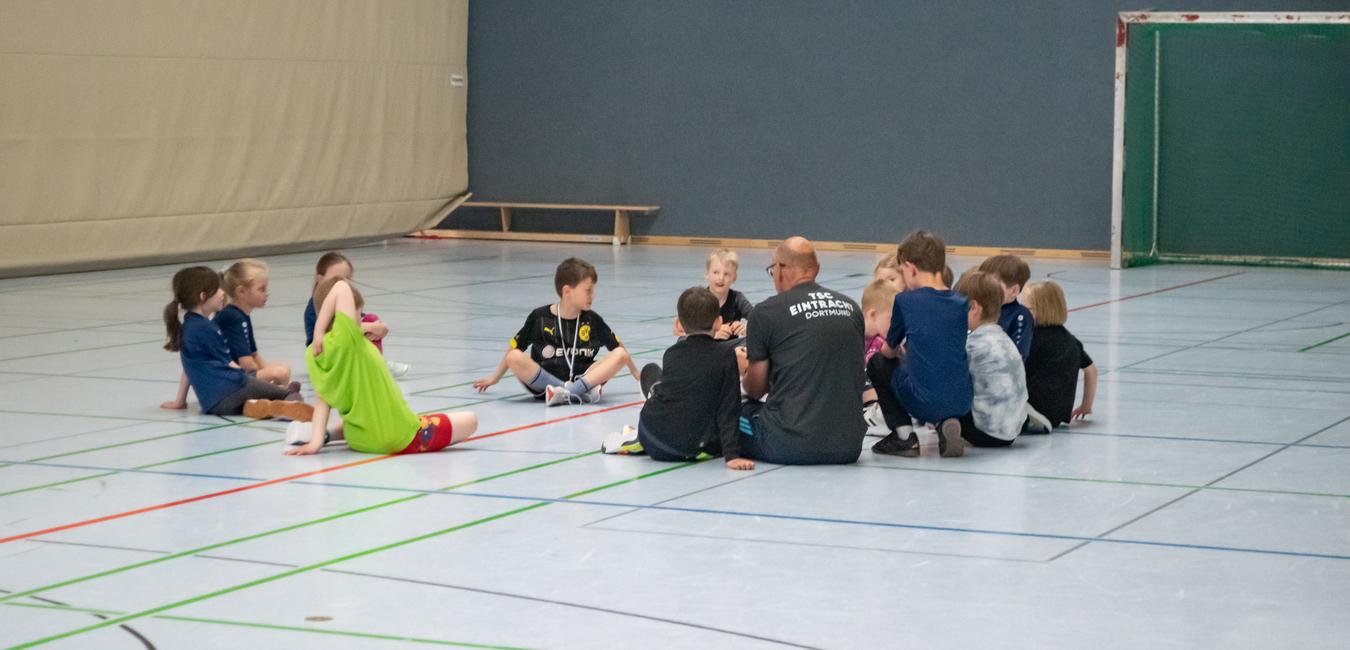 Gruppe von Kindern sitzt im Kreis auf einem Sportboden, um einen Erwachsenen, der ihnen etwas erklärt.