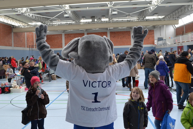 Maskottchen Victor in einem Sporthalle, umgeben von Menschen bei einem Flohmarkt. Es hebt die Arme zur Feier.