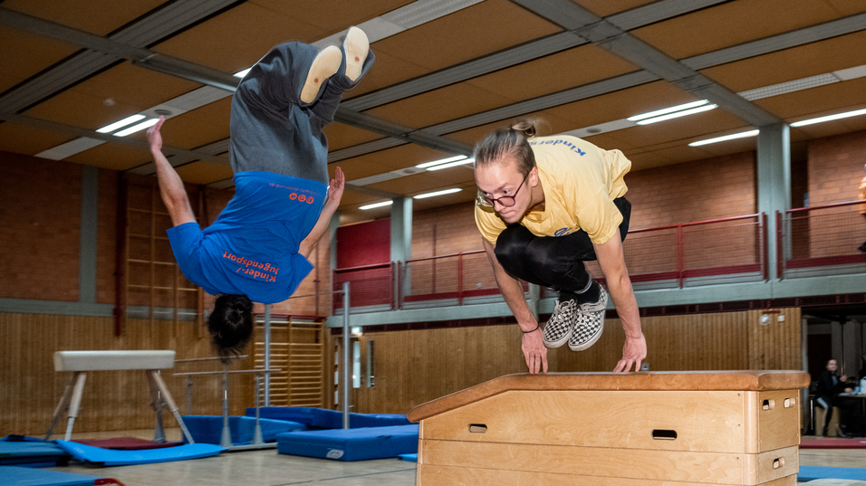 Zwei Athleten springen akrobatisch &uuml;ber eine Holzbank in einer Sporthalle mit &Uuml;bungsger&auml;ten im Hintergrund.