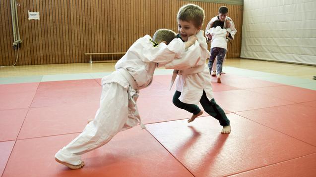 Zwei Kinder im wei&szlig;en Judo-Anzug &uuml;ben in der Sporthalle, w&auml;hrend im Hintergrund weitere Teilnehmer trainieren.