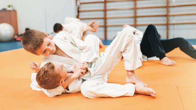 Zwei Kinder ringen auf einer orangefarbenen Mattenfläche in Judoanzügen. Im Hintergrund sind weitere Kinder aktiv.