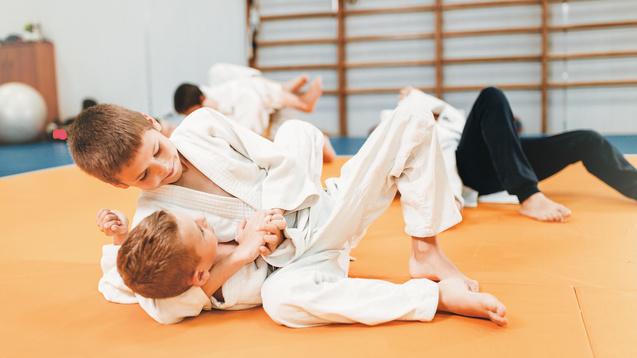 Zwei Kinder ringen auf einer orangefarbenen Mattenfläche in Judoanzügen. Im Hintergrund sind weitere Kinder aktiv.
