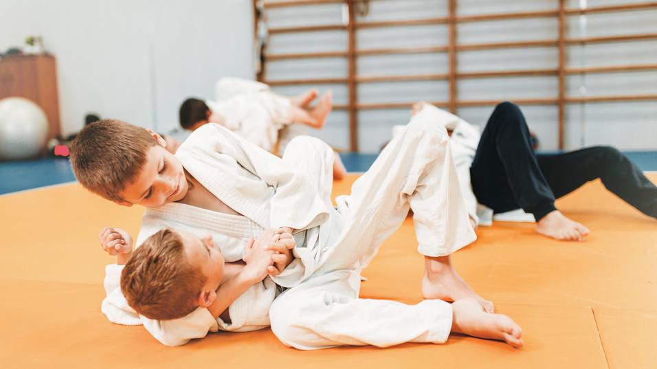 Zwei Kinder ringen auf einer orangefarbenen Mattenfläche in Judoanzügen. Im Hintergrund sind weitere Kinder aktiv.