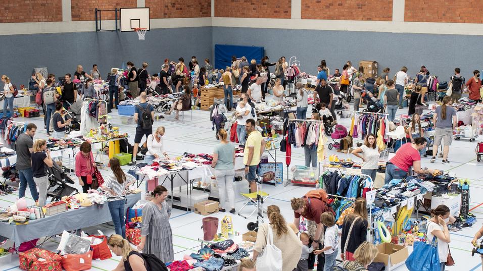 Blick in eine große Sporthalle mit vielen Menschen und Verkaufsständen, gefüllt mit Kleidung und Spielzeug.