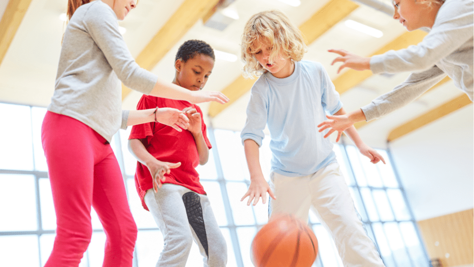 Vier Kinder spielen in einer sportlichen Halle mit einem Basketball, während sie den Ball anvisieren.