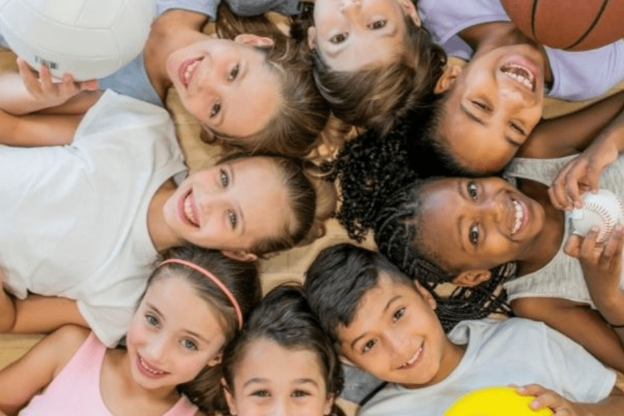 Gruppe von Kindern mit verschiedenen Sportb&auml;llen, die l&auml;chelnd auf einem Parkettboden liegen. Text: &bdquo;Kindersport Sporties 2024/2025&ldquo;.
