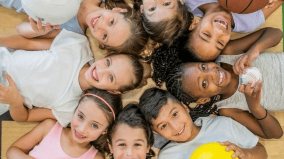 Gruppe von Kindern mit verschiedenen Sportbällen, die lächelnd auf einem Parkettboden liegen. Text: „Kindersport Sporties 2024/2025“.