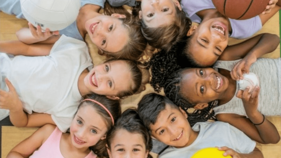 Gruppe von Kindern mit verschiedenen Sportbällen, die lächelnd auf einem Parkettboden liegen. Text: „Kindersport Sporties 2024/2025“.