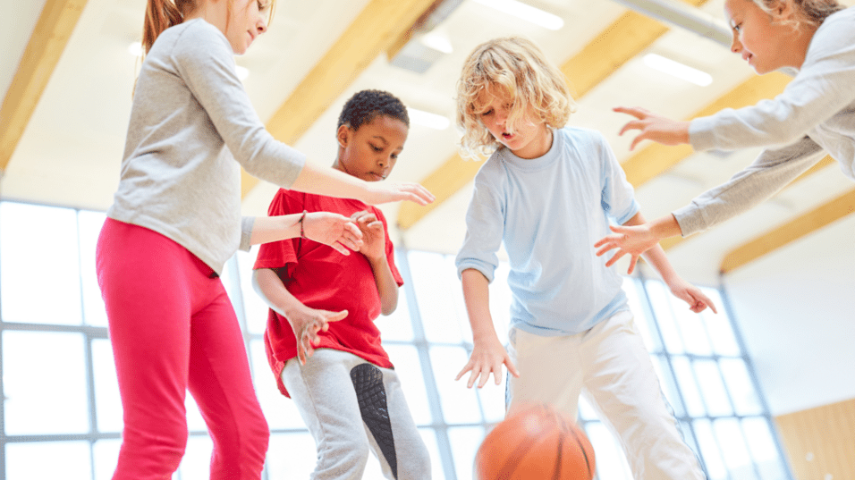 Vier Kinder spielen in einer sportlichen Halle mit einem Basketball, während sie den Ball anvisieren.