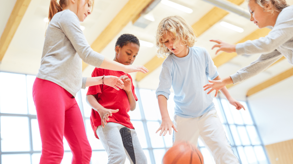 Vier Kinder spielen in einer sportlichen Halle mit einem Basketball, während sie den Ball anvisieren.