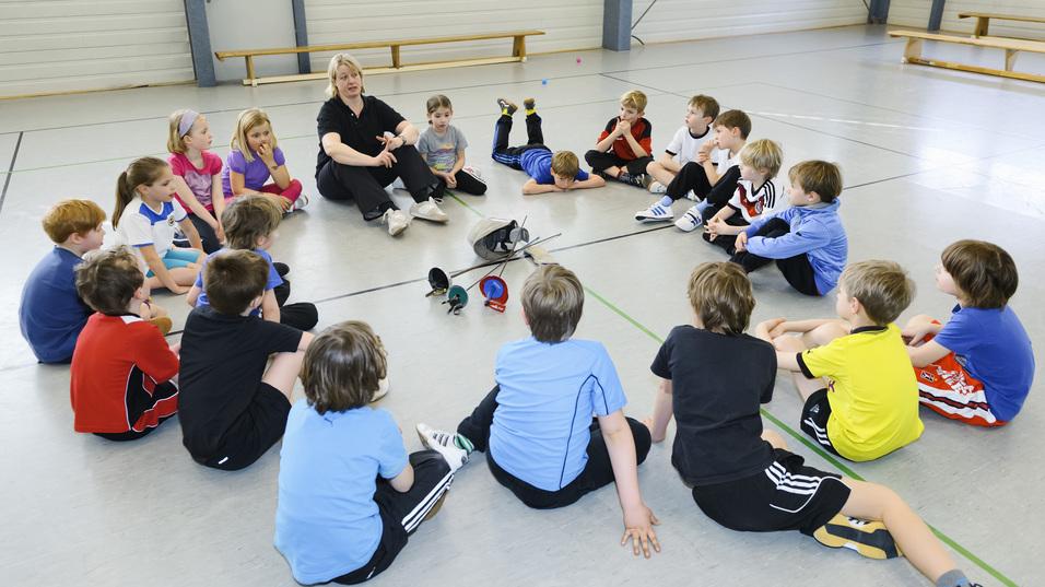 Gruppierung von Kindern in Sportkleidung, die einem Trainer in einer Halle zuhören. Sportmaterialien liegen auf dem Boden.