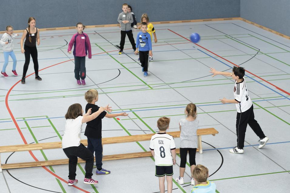 Gruppe von Kindern spielt mit einem blauen Ball in einer Turnhalle, einige stehen und andere werfen.
