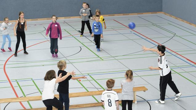 Gruppe von Kindern spielt mit einem blauen Ball in einer Turnhalle, einige stehen und andere werfen.