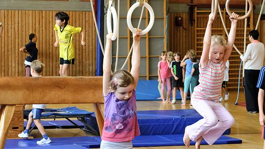 Kinder turnen in einer Sporthalle: einige balancieren auf einem Schwebebalken, andere benutzen Ringe oder Kasten.