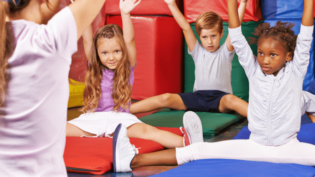 Gruppen von Kindern in sportlicher Kleidung sitzen auf Matten und heben die H&auml;nde. Eine Trainerin steht vor ihnen.