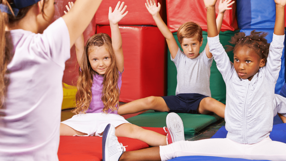 Gruppen von Kindern in sportlicher Kleidung sitzen auf Matten und heben die Hände. Eine Trainerin steht vor ihnen.