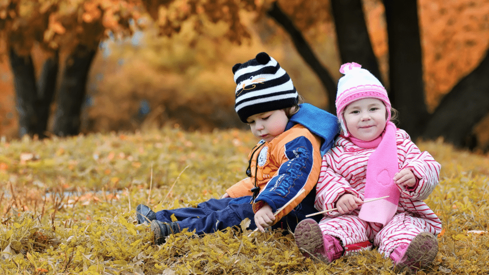 Zwei kleine Kinder sitzen auf buntem Laub in einem Herbstgarten, eines tr&auml;gt eine M&uuml;tze mit Streifen und das andere eine M&uuml;tze mit Bommel.