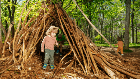 Kinder spielen in einem selbstgebauten H&uuml;ttchen aus Zweigen im Wald. Gr&uuml;ne B&auml;ume und Laub umgeben die Szene.