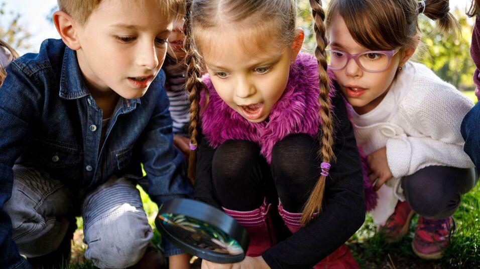 Vier Kinder beobachten gespannt durch eine Lupe, w&auml;hrend sie auf dem Gras sitzen und sich umschauen.