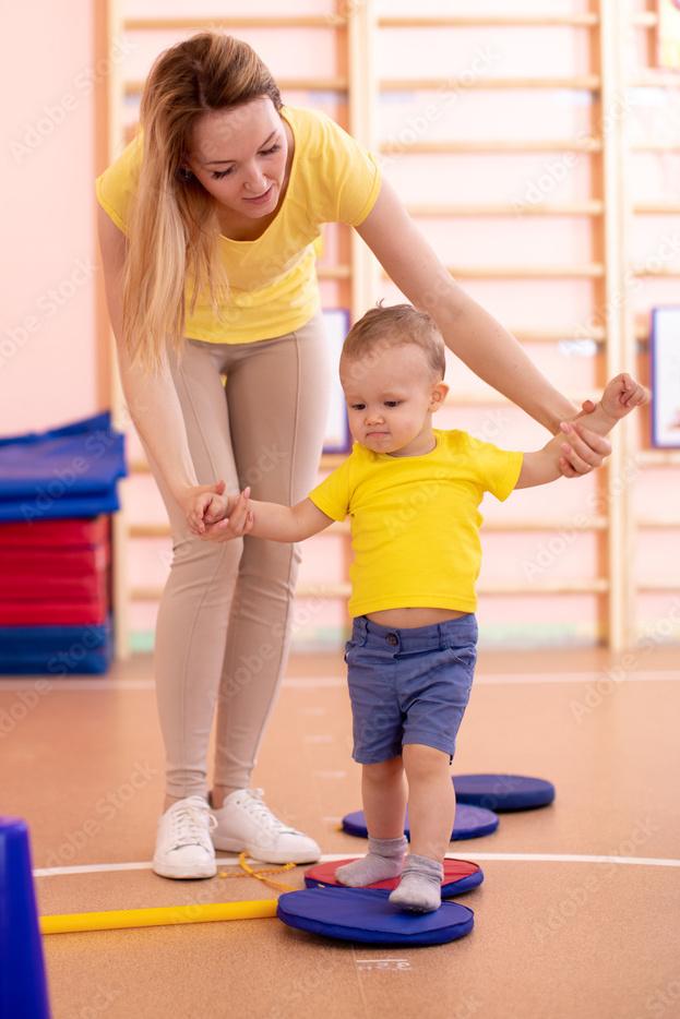 Eine Frau h&auml;lt die H&auml;nde eines kleinen Jungen, der auf einem blauen Gleichgewichtsspielger&auml;t in einem Turnraum steht.