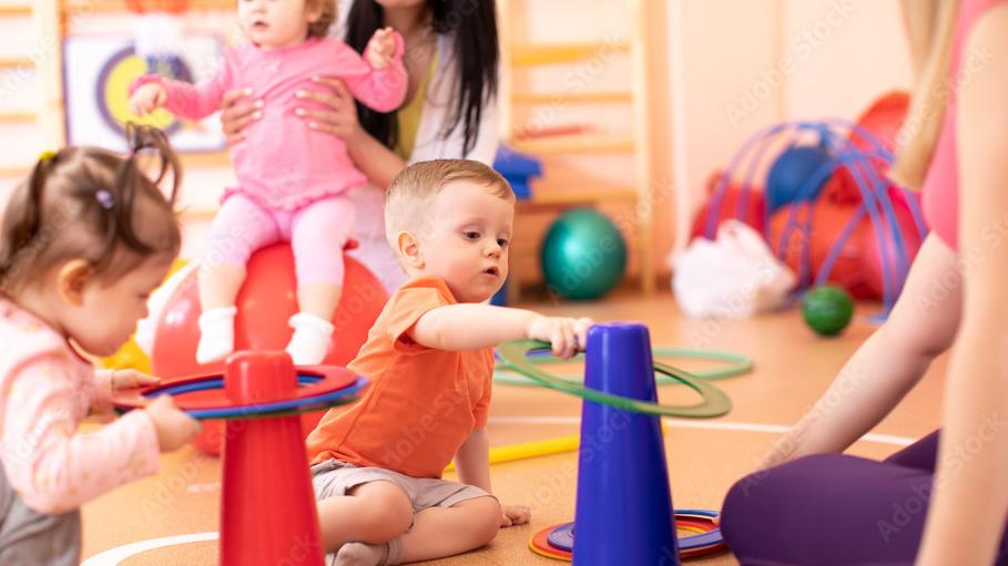 Kinder spielen in einem Gruppenraum mit bunten Stapelringen und k&uuml;mmern sich umeinander.