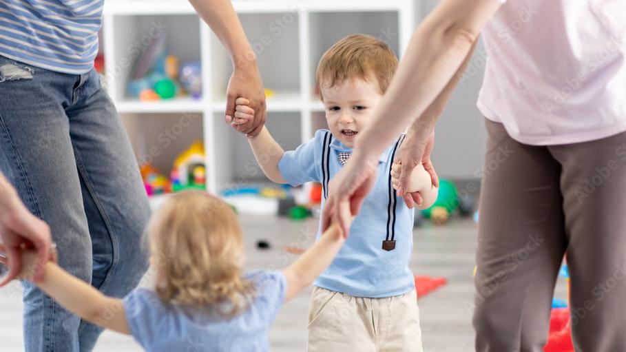 Familie h&auml;lt H&auml;nde beim Spielen auf einem bunten Spielteppich mit Spielsachen im Hintergrund.