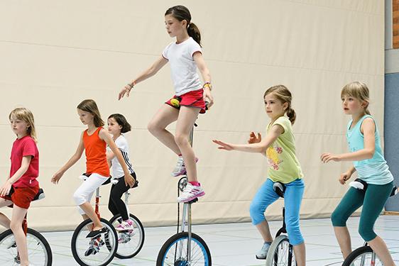 Gruppe von M&auml;dchen f&auml;hrt auf Einr&auml;dern in einer Sporthalle, einige balancieren und zeigen verschiedene Fahrtechniken.