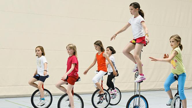 Gruppe von Mädchen fährt auf Einrädern in einer Sporthalle, einige balancieren und zeigen verschiedene Fahrtechniken.