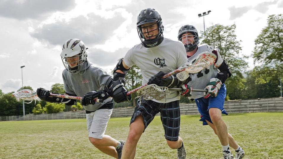 Drei Lacrosse-Spieler sprinten &uuml;ber ein gr&uuml;nes Spielfeld, ausgestattet mit Helmen und Lacrosse-Schl&auml;gern.