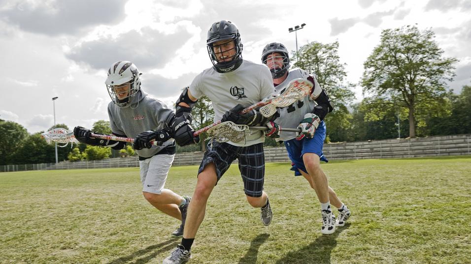 Drei Lacrosse-Spieler sprinten &uuml;ber ein gr&uuml;nes Spielfeld, ausgestattet mit Helmen und Lacrosse-Schl&auml;gern.