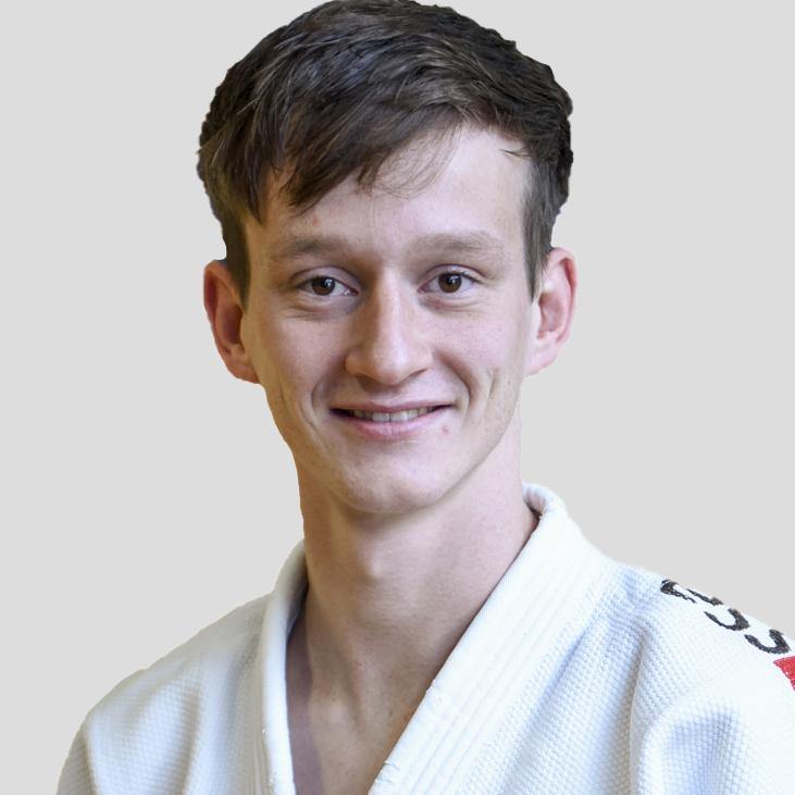 Junger Mann lächelt in Judo-Anzug mit weißem Gi vor grauem Hintergrund.