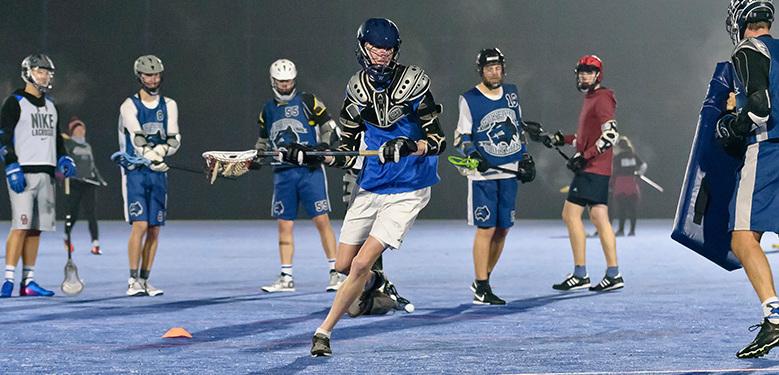 Lacrosse-Spieler trainieren nachts auf einem blauen Platz, einige mit Schlägern und Schutzausrüstung.