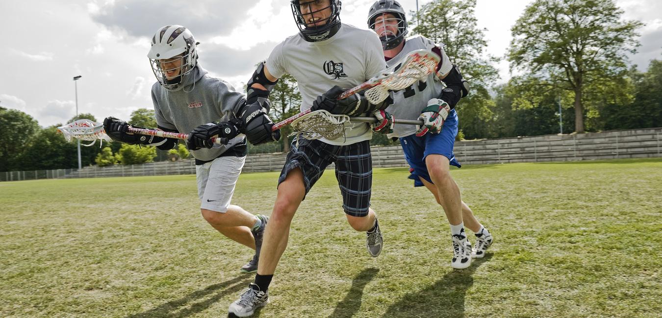 Drei Lacrosse-Spieler sprinten über ein grünes Spielfeld, ausgestattet mit Helmen und Lacrosse-Schlägern.
