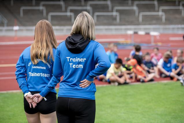 Zwei Trainerinnen in blauen Sweatshirts stehen vor einer Gruppe von Kindern auf einer Sportanlage.