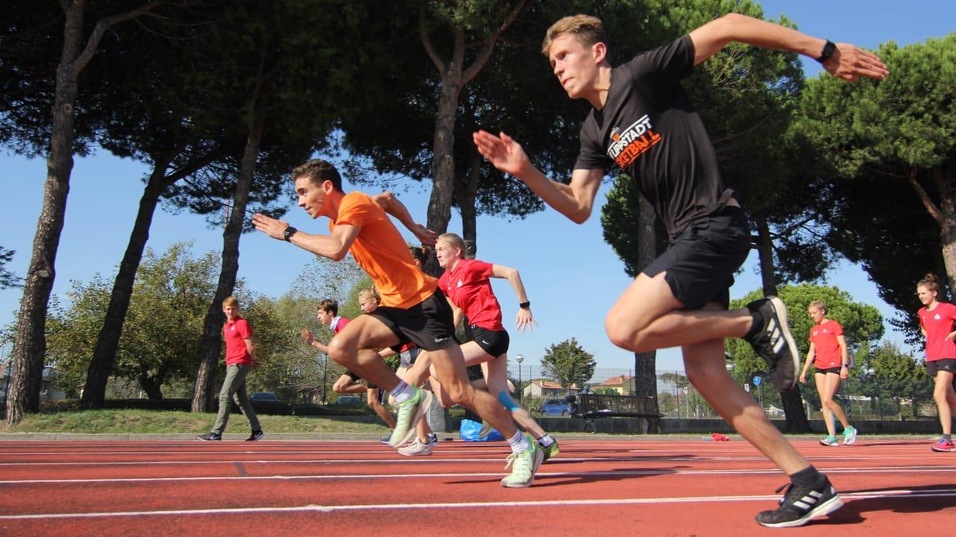 Sechs Athleten sprinten auf einer roten Laufbahn unter blauem Himmel und begr&uuml;nten B&auml;umen.