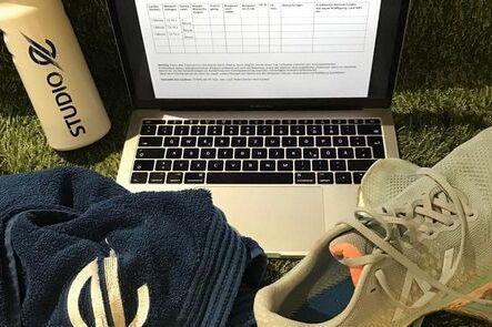 Laptop mit offener Tabelle, eine Wasserflasche, ein Handtuch und Laufschuhe auf gr&uuml;nem Untergrund.