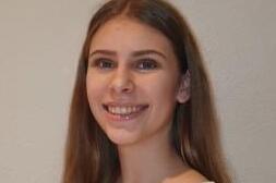 Junge Frau mit langen, braunen Haaren, l&auml;chelt vor grauer Wand. Sie tr&auml;gt ein helles, schlichtes Oberteil.